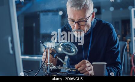Electronics Maintenance Engineer Enters His Workshop with Coffee Beginss Soldering of Circuit Board, Looking through Magnifying Glass, Consults Stock Photo