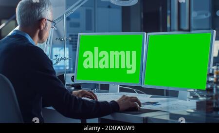 Professional Engineer Works on Computer with Two Green Mock-up Screen Displays. In the Background Industrial Electronics Design Agency. Over the Stock Photo