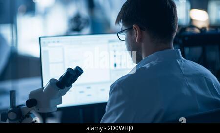 Shot of a Male IT Scientist Uses Computer with Electronic Microscope. In the Background Technology Development Laboratory with Scientists, Engineers Stock Photo