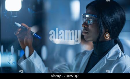 Close-up Shot of an Smart and Beautiful African American Female Scientist Wearing White Coat and Protective Glasses Writes Formula on Glass Whiteboard Stock Photo