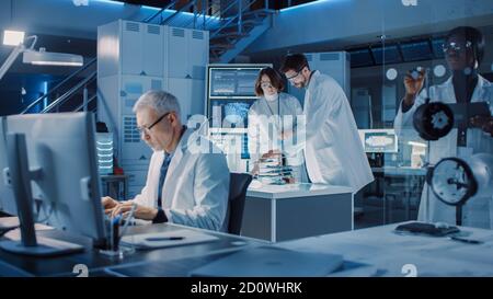 Diverse International Team of Industrial Scientists and Engineers Wearing White Coats Working on Heavy Machinery Design in Research Laboratory Stock Photo