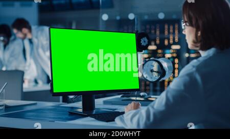 Over the Shoulder Shot: Female IT Scientist Uses Green Mock-up Screen Computer. In the Background Technology Research And Development Laboratory with Stock Photo