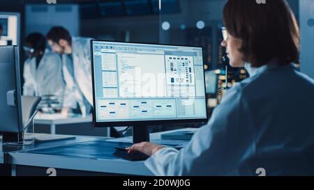 Over the Shoulder Shot: Female IT Scientist Uses Computer Showing System Monitoring and Controlling Program. In the Background Technology Development Stock Photo