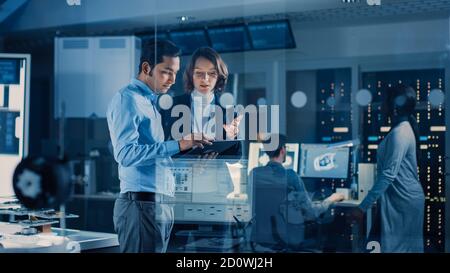 In Technology Research Facility: Female Project Manager Talks With Chief Engineer, they Consult Tablet Computer. Team of Industrial Engineers Stock Photo