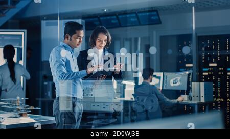 In Technology Research Facility: Female Project Manager Talks With Chief Engineer, they Consult Tablet Computer. Team of Industrial Engineers Stock Photo