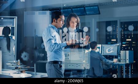 In Technology Research Facility: Female Project Manager Talks With Chief Engineer, they Consult Tablet Computer. Team of Industrial Engineers Stock Photo