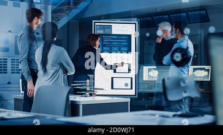 Engineers Meeting in Technology Research Laboratory: Female Engineer Leads Presentation Writes on Interactive Digital Whiteboard, Shows Machine Stock Photo