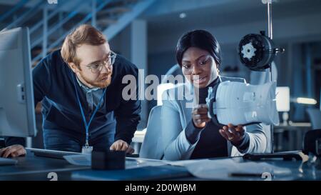 Electronics Development Engineer Working at His Desk, Talks with Project Manager, Shows Mechanism Prototype Construction. Team of Professionals Stock Photo