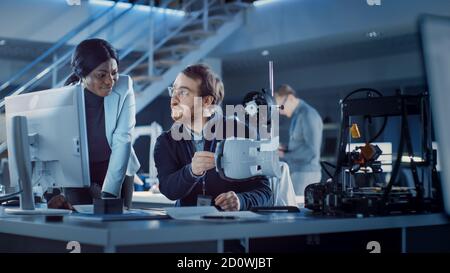 Electronics Development Engineer Working at His Desk, Talks with Project Manager, Shows High Tech Prototype Component. Team of Professionals Working Stock Photo