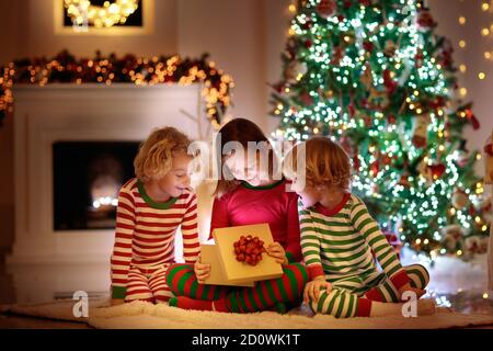 Children at Christmas tree and fireplace on Xmas eve. Family with kids celebrating Christmas at home. Boy and girl in matching pajamas decorating xmas Stock Photo