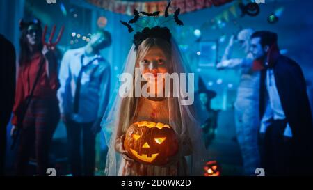 Halloween Costume Party: Smiling Little Girl in a Bloody White Bride Dress Holding Burning Pumpkin Head. Zombie, Blood Thirsty Dracula, Mummy Stock Photo