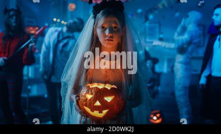 Halloween Costume Party: Little Girl in a Bloody White Bride Dress Holding Scary Doll. Zombie, Blood Thirsty Dracula, Mummy, Bewitching Witch and Stock Photo