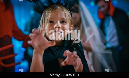 Halloween Costume Party: Cute Little Bat Girl with Sharp Fangs Makes Scary Funny Faces. In the Background Monster Party in Decorated Room and Disco Stock Photo