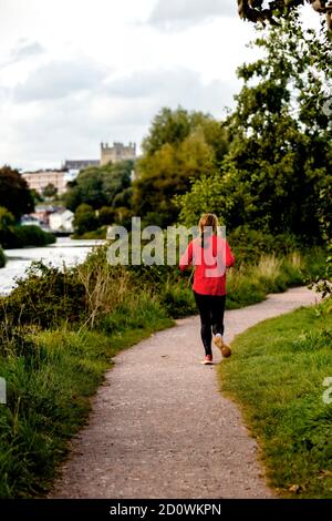 Young blond Woman jogging on a green footpath along a river in Autumn seen from the back Stock Photo