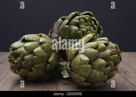 Three fresh organic purple artichokes with stem on a rustic wooden surface and dark background Stock Photo