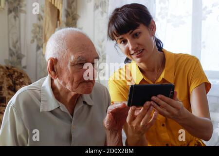 a man of the 90s with an adult granddaughter, sitting together on a cozy sofa with a phone in his hands, looking at photos. a woman in a yellow dress Stock Photo