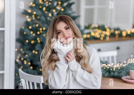 Beautiful curly blonde with long hair by the Christmas trees with garlands of lights. Close-up portrait of a Young woman with a white smile in a gray Stock Photo