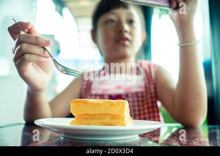 Asian little child girl eating chiffon cake with looking smart phone Stock Photo