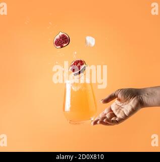 Woman hand support fly glass of pink cocktail drink with splash, juice slice fig falling in glass. Summer art food concept on peach color background Stock Photo