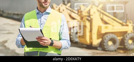 Young civil engineer wearing helmet writing report while standing at construction site Stock Photo
