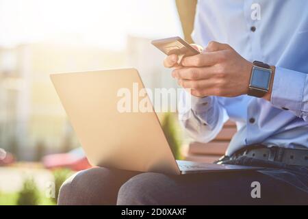 Online business. Cropped shot of a businessman using smartphone and laptop while sitting on the bench outdoors and working remotely Stock Photo
