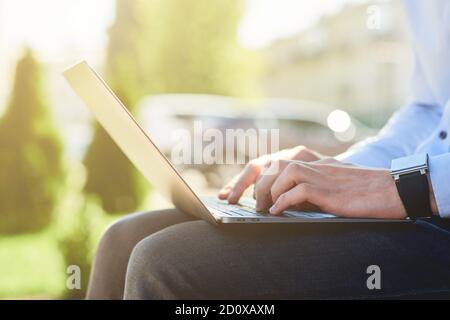 Cropped shot of a male freelancer working online on laptop while sitting on the bench outdoors on a sunny day Stock Photo
