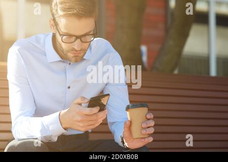 Business message. Focused businessman using smartphone and drinking coffee while sitting on the bench outdoors Stock Photo