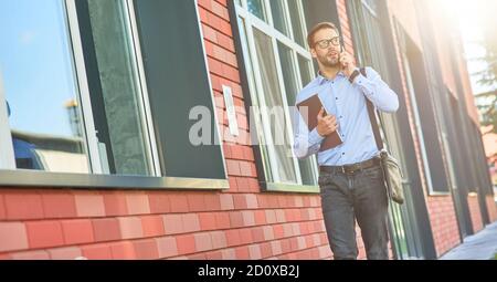 Business talk. Young confident caucasian businessman wearing blue shirt and eyeglasses holding his laptop and talking by phone while walking outdoors Stock Photo