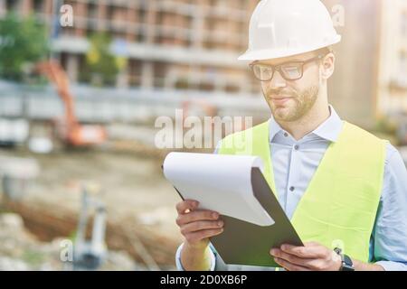 Controlling the project. Young focused civil engineer or construction supervisor wearing helmet reading report while inspecting construction site Stock Photo