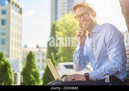 Good news. Young happy businessman talking by phone and and smiling while sitting on the bench outdoors and working online, using laptop Stock Photo