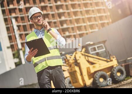 Important call. Young civil engineer or construction supervisor wearing helmet talking by phone while inspecting building site Stock Photo