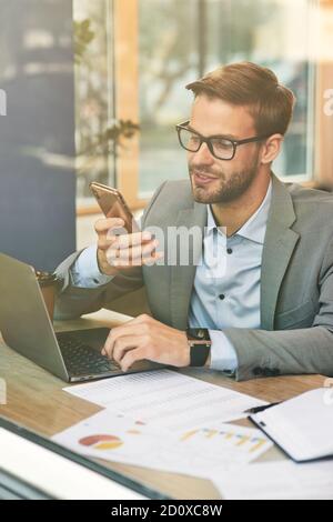 Vertical shot of a young focused businessman or male freelancer using his smartphone while working online in cafe. Laptop and documents on wooden Stock Photo