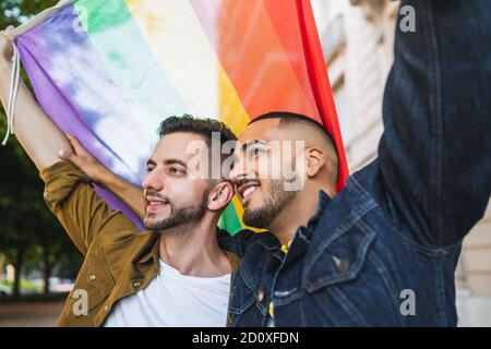 Portrait of young gay couple embracing and showing their love with rainbow flag at the street. LGBT and love concept. Stock Photo