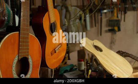 Two hanging wooden guitars and guitar parts on the carpentry table in a workshop with wall full of tools in the background Stock Photo