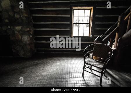 Empty Room With Single Chair. Sunlight streams through the window of a cabin with an empty wooden chair in the corner Stock Photo