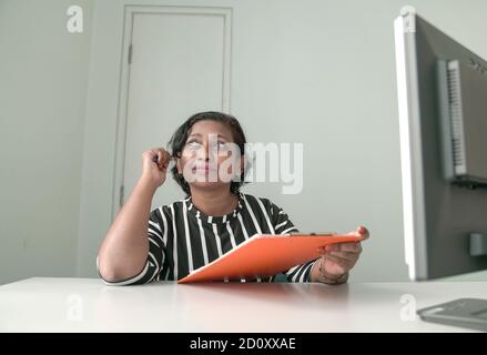 Woman at work, in front of computer, holding a file, with a thinking expression Stock Photo
