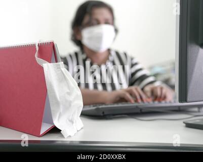 Face mask hang on a table calendar at the work place, with blur background of businesswoman typing. Stock Photo