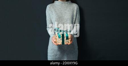 Woman in gray pullover holding luminous gift box in hands on dark background. Glowing yellow light bulbs into transparent gift box with dark green ribbon. Christmas and New Year concept Stock Photo