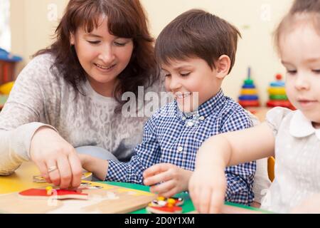 beautiful mature caucasian female teacher collects the puzzle with a pupil,  a boy of preschool age. In the classroom, at the green table. Stock Photo