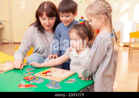 beautiful mature woman teacher collect puzzle with three pupils of a kindergarten classroom at the green table. Horizontal color image. Stock Photo