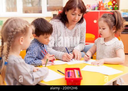 Kindergarten teacher, beautiful, mature caucasian woman, brown hair, teaches pupils drawing. Two little girls and a boy. Stock Photo