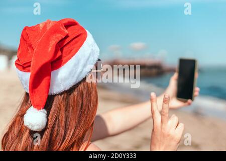 Young beautiful girl in a red santa claus hat and swimsuit makes a selfie on the seashore Stock Photo