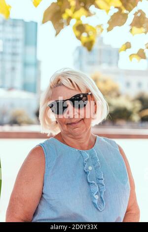 Portrait of adult blonde woman in sunglasses smiling at camera outdoors in park Stock Photo