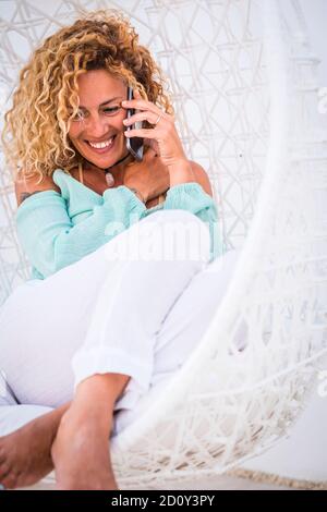 Cheerful beautiful adult young caucasian woman portrait have a phone call sitting on a white chairs at home - concept of luxury and rich people lifest Stock Photo