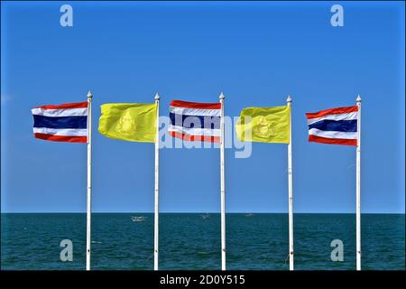 A row of Thai flags and the yellow King’s flag flapping in a strong onshore wind against a background of blue sky and sea. Stock Photo