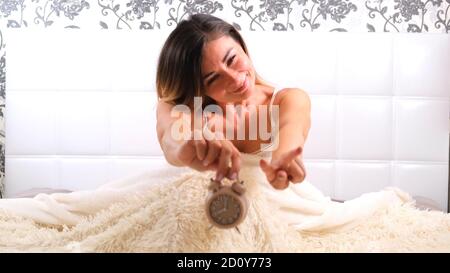 female rises in bed with alarm clock, enjoys the morning. The concept of starting a new day, relaxing on the weekend, no need to rush anywhere, good Stock Photo