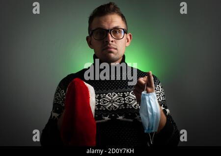 The man in glasses choosing between the medical mask and a Santa hat, the concept of pandemic for Christmas, Stock Photo