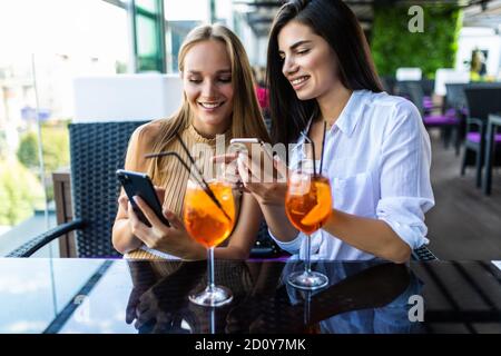 Two women smiling and using cellphones while drinking cocktails at table in cafe outdoors Stock Photo