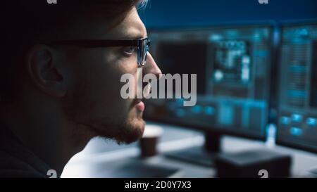Portrait of IT Specialist Wearing Glasses Works on Personal Computer with Screens Showing Software Program with Coding Language Interface. Dark Colors Stock Photo