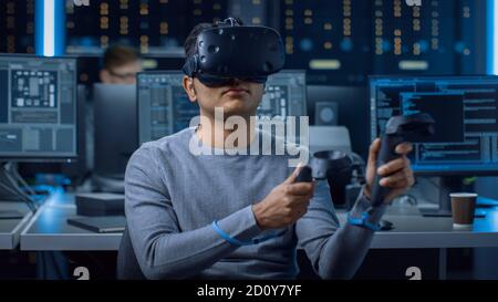 Portrait of Software Delevoper Wearing Virtual Reality Headset Using Controllers to Develop and Program VR Gaming Applications. In Background Stock Photo